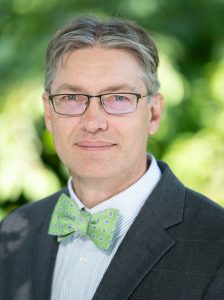 Eric Meyers is a man with light skin tone. His short grey hair is worn in a middle part. He is wearing black rectangular thin framed glasses, a lime green bowtie, and a white dress shirt with a black blazer. He is smiling with his mouth closed at the camera.