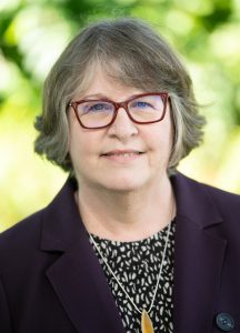 Luanne Sinnamon is a woman with light skin tone and grey hair that is cut into a bob with curtain bangs in a side part. She is wearing red rectangular glasses, a dark purple blazer, and a black top with small irregular shapes in white as the pattern. She is smiling directly at the camera. The background is green leaves blurred out in a bokeh effect.