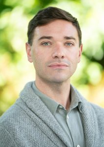 Cameron is a man with light skin tone and short, dark brown hair that he combs to the side. His body is angled to the right, but his head is turned to face the camera. at the camera. He is wearing a green-grey button up, with a grey cardigan overtop.