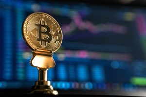 Gold Bitcoin in foreground, with blurry computer background.